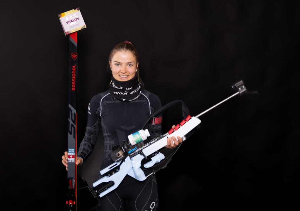 Eine Sportlerin in schwarzem Sportoutfit hält Langlaufski und ein Biathlongewehr in der Hand und steht vor einem schlichten schwarzen Hintergrund, bereit für ihre nächste Herausforderung.