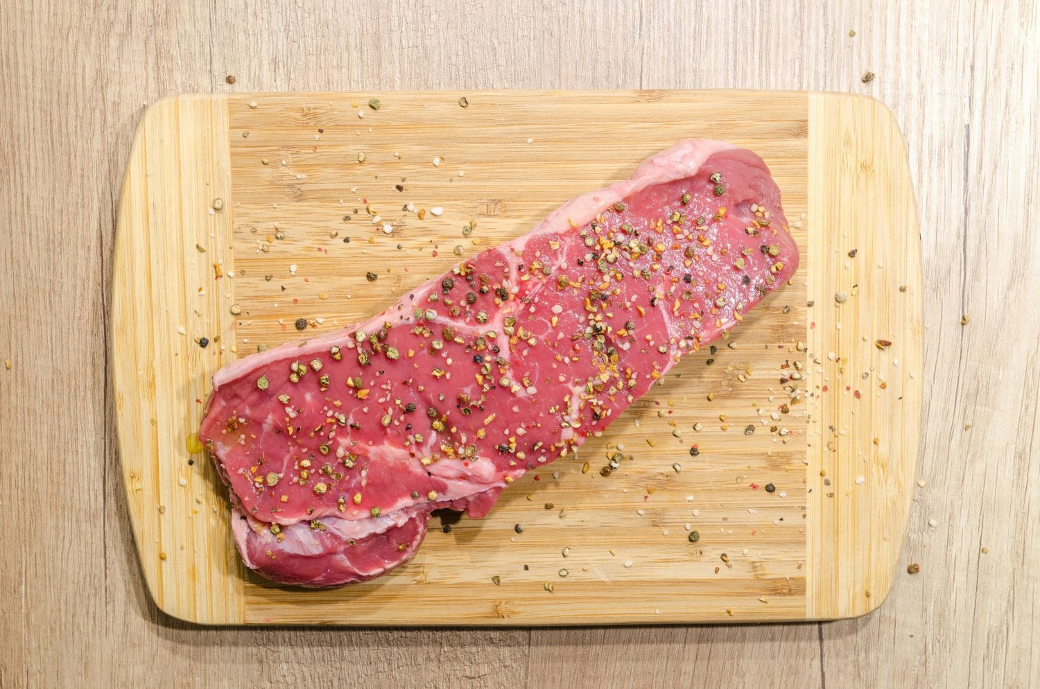 Ein rohes Steak, bedeckt mit gemischten Pfefferkörnern und Salz, liegt auf einem Holzschneidebrett auf einem hellen Holztisch und ist somit eine proteinreiche Mahlzeit und eine natürliche Zinkquelle, ideal als Ergänzung zu Ihrer täglichen Nahrungsergänzung.