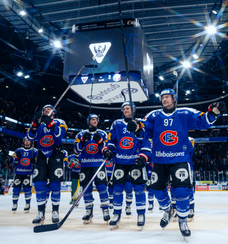 Eine Gruppe Eishockeyspieler in blauen Trikots steht auf dem Eis, reckt ihre Schläger in die Höhe und winkt der Menge in der hell erleuchteten Arena zu. Auf der großen Leinwand über ihnen sind das Teamlogo, der Spielstand und Sponsoren zu sehen, die für Vitalstoffe werben.