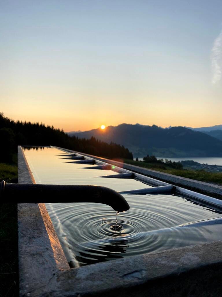 Ein metallener Wasserspeier tropft in eine lange, reflektierende Rinne und erzeugt Wellen. Der Hintergrund zeigt einen malerischen Sonnenuntergang über Bergen und einem entfernten See, am Horizont zeichnen sich Bäume und Natur ab.