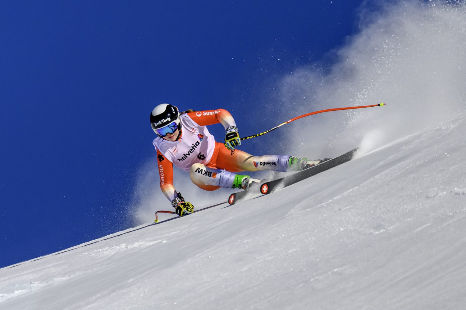Allegra Frei, in einem rot-weißen Rennanzug und mit Helm, rast einen verschneiten Hang hinunter und neigt sich dabei stark, während hinter ihr der Schnee gegen den klaren blauen Himmel spritzt.