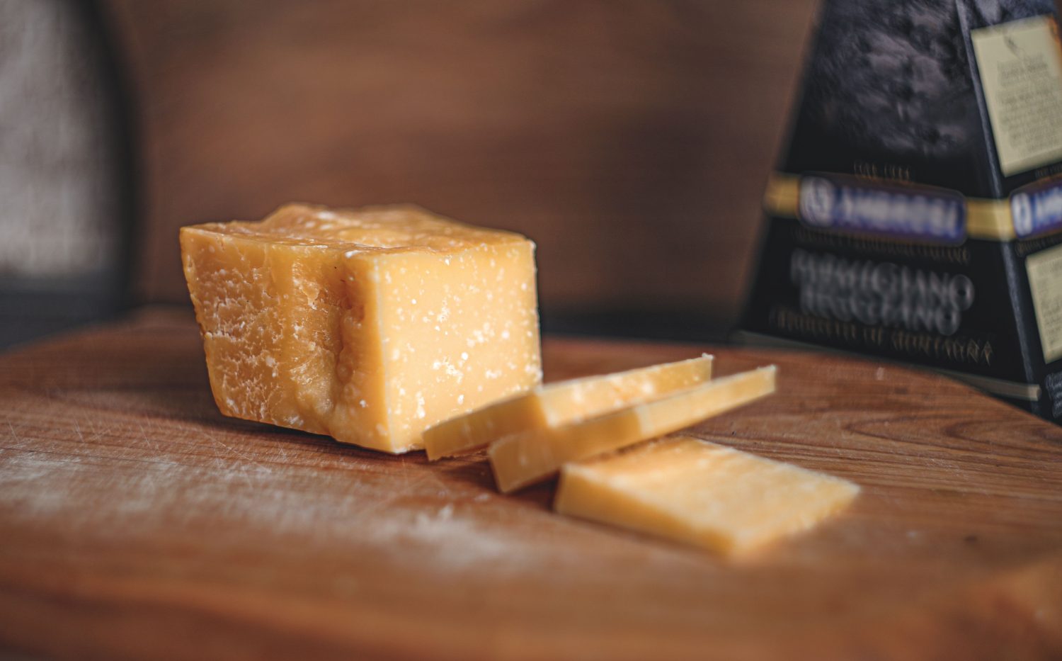 Ein Stück Hartkäse mit drei dünnen Scheiben auf einem Holzschneidebrett, im Hintergrund ein Käsekeil und eine verschwommene Verpackung – perfekt für alle, die auf ihre Ernährung achten oder Symptome im Zusammenhang mit einer Histaminintoleranz in den Griff bekommen möchten.
