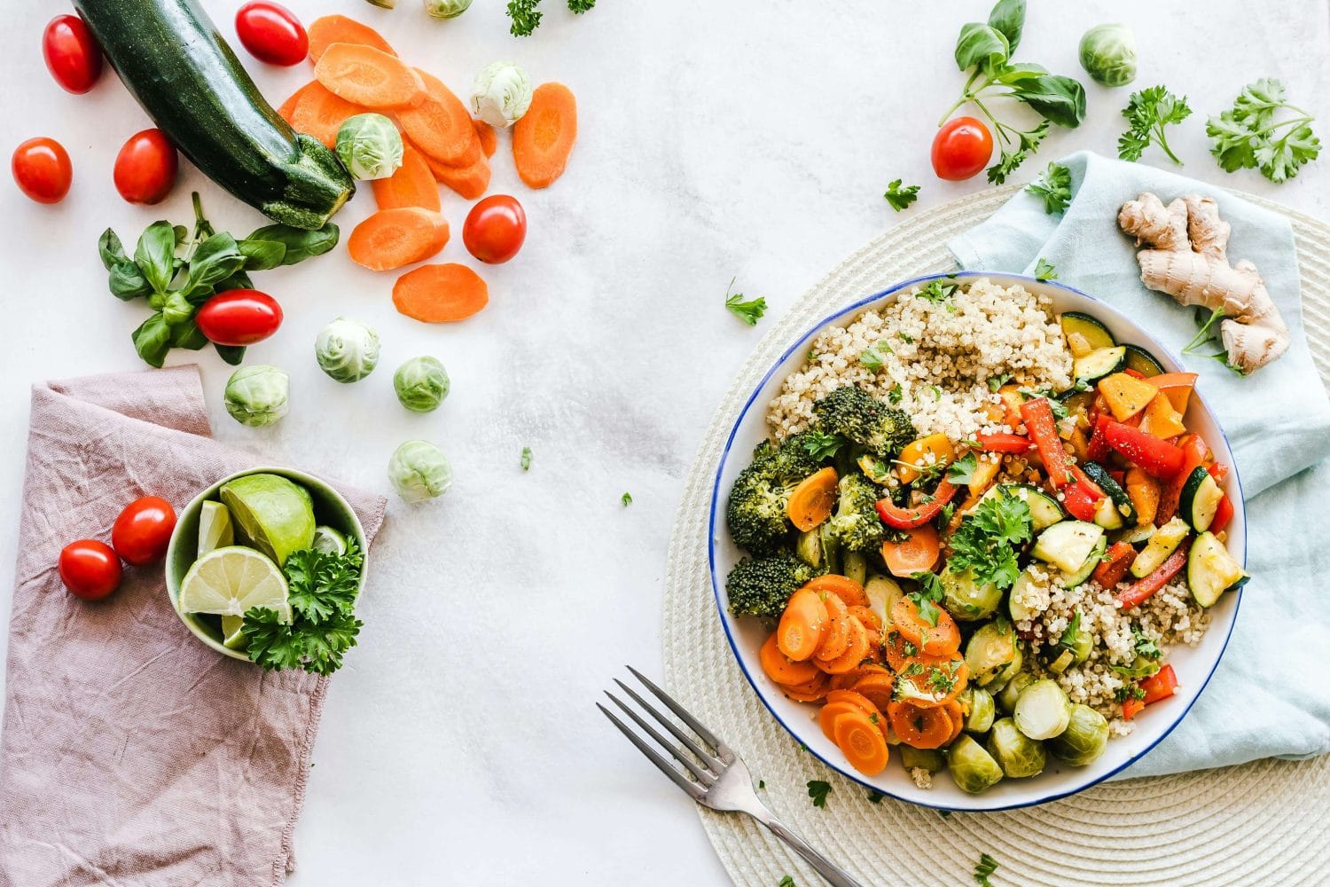 Eine farbenfrohe Schüssel mit gekochtem Gemüse und Quinoa steht auf einem Tischset, umgeben von frischen Zutaten wie Zucchini, Karotten, Tomaten, Rosenkohl, Ingwer und Limette auf einem weißen Tisch. Eine nahrhafte Mahlzeit erwartet Sie neben einer Gabel.