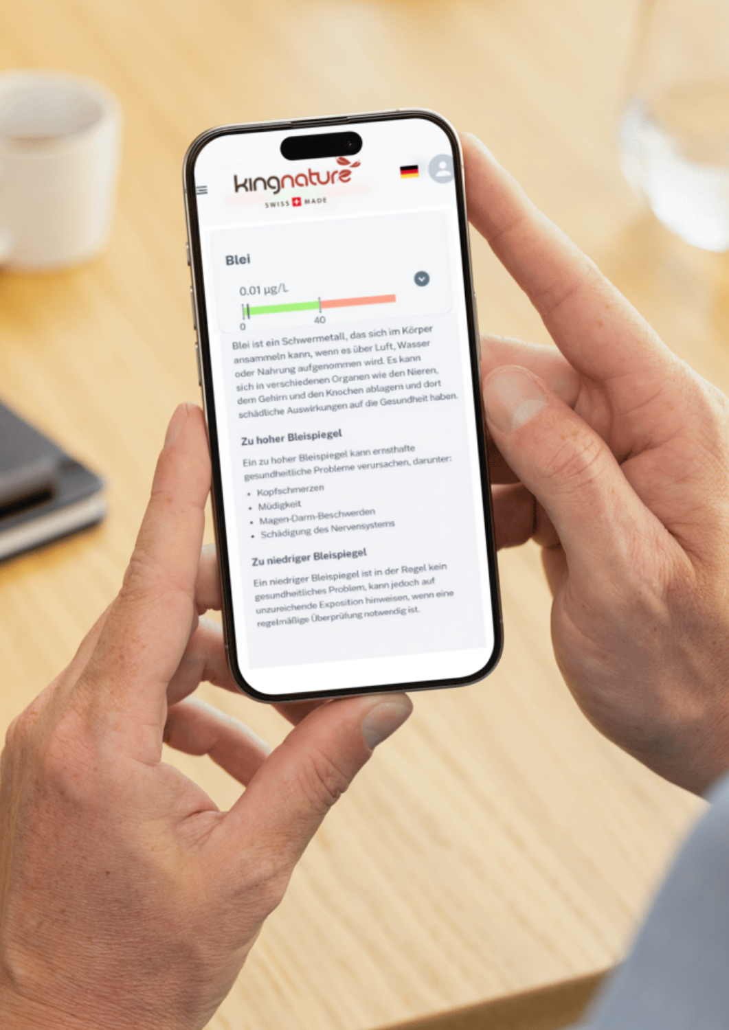 Eine Person hält ein Smartphone in der Hand, auf dem eine Webseite über Bleigehalt messen mit einem Messwert von 0,01 μg/L angezeigt wird, während sie an einem Holztisch mit einem Notebook und einem Glas Wasser daneben sitzt.