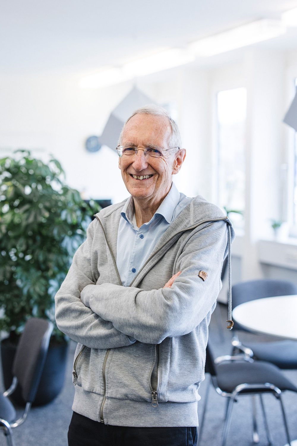 Ein älterer Mann mit Brille und hellblauem Hemd unter einem grauen Kapuzenpullover mit Reißverschluss steht lächelnd und mit verschränkten Armen in einem hellen kingnature-Büro – einer der Helden aus dem Verwaltungsrat, umgeben von Pflanzen, Stühlen und Tischen.