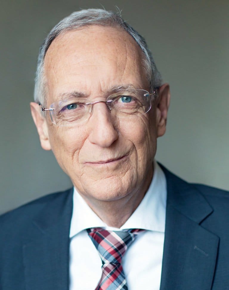 Ein älterer Mann mit kurzen grauen Haaren, Brille und hellen Augen trägt einen dunklen Anzug, ein weißes Hemd und eine gemusterte Krawatte. Sein direkter Blick in die Kamera lässt vermuten, dass er bereit für ein Interview ist – vielleicht ist es Dr. med. Lüscher selbst.