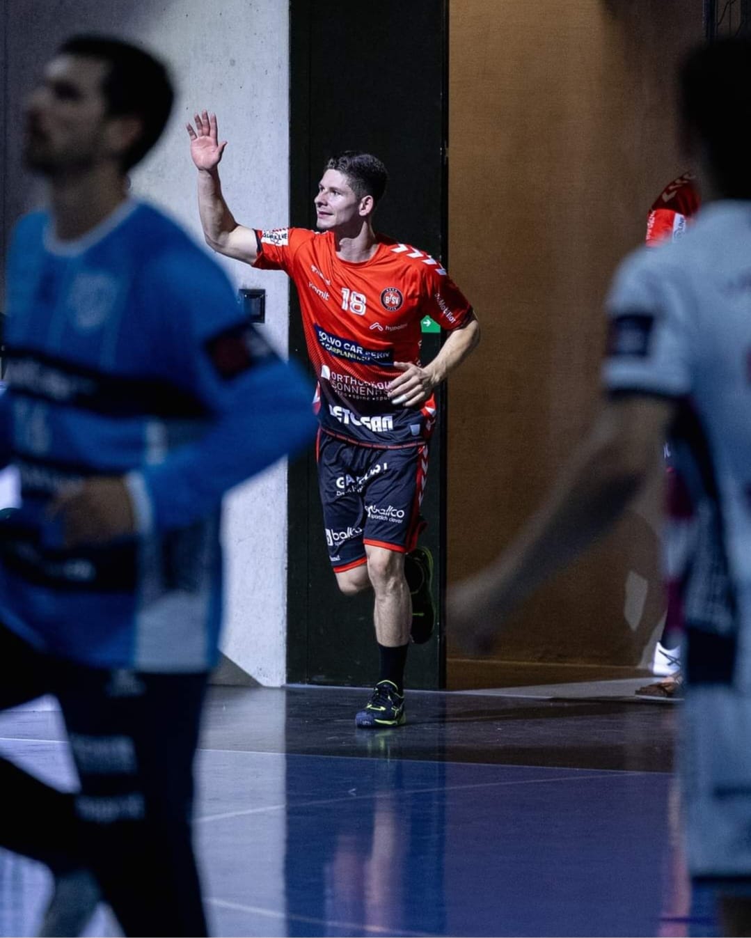 A male handball player in a red jersey, numbered 18, runs out from a doorway onto the court, smiling and waving. Other players in blue and white jerseys are visible in the foreground.