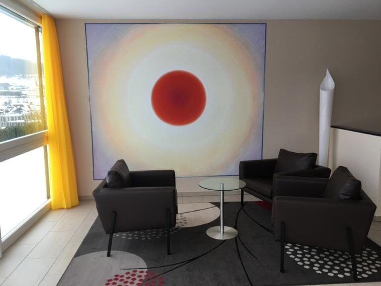 Eine moderne Sitzecke mit zwei schwarzen Sesseln, einem Couchtisch aus Glas und einem dunklen Teppich. Dahinter sticht Alex Peter Zwikers großes abstraktes Gemälde eines roten Kreises mit strahlenförmigen Ringen hervor. Gelbe Vorhänge rahmen das Fenster links ein und schaffen eine Atmosphäre des „Ganzheitlichen Coachings“.