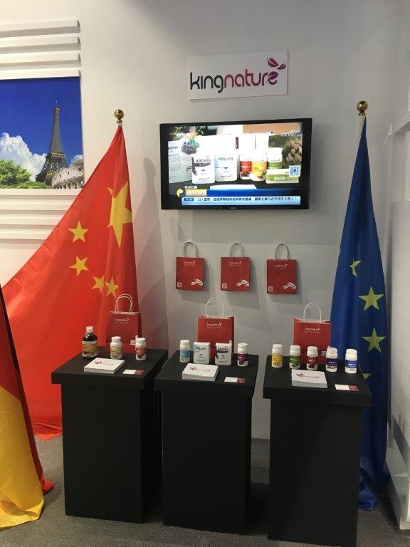 Präsentation der kingnature-Nahrungsergänzungsmittel auf drei schwarzen Ständen, dahinter die Flaggen von China, Deutschland und der EU. Rote Geschenktüten und ein Fernsehbildschirm mit kingnature-Produkten unterstreichen die Präsenz des Unternehmens auf der Nordostasien-Expo.