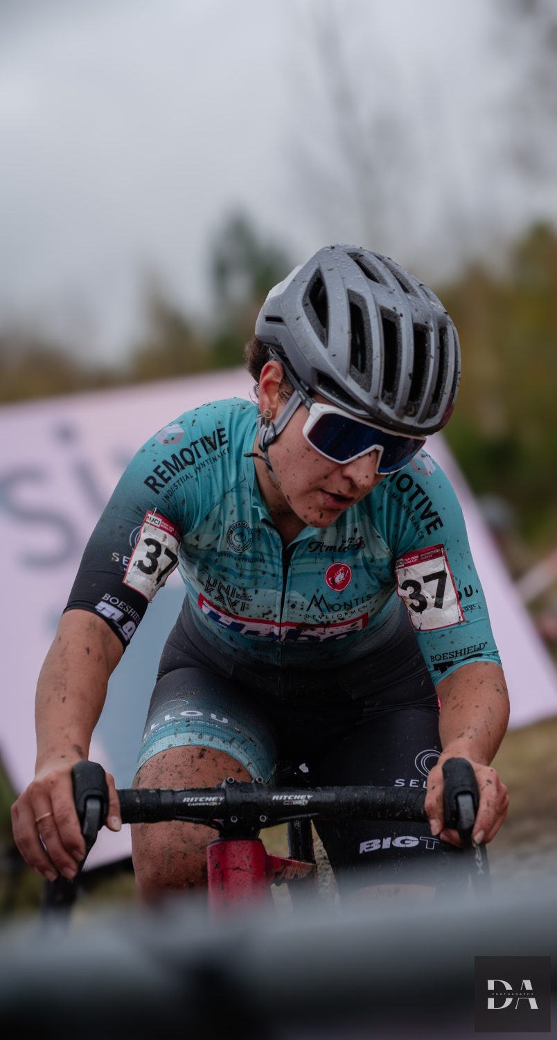 Radfahrerin Zina Barhoumi trägt ein blaugrün-schwarzes Rennoutfit, Helm und Sonnenbrille und ist während eines Radrennens mit Schlamm bedeckt. Vor einem unscharfen Hintergrund ist die Nummer 37 auf ihren Armen zu sehen.