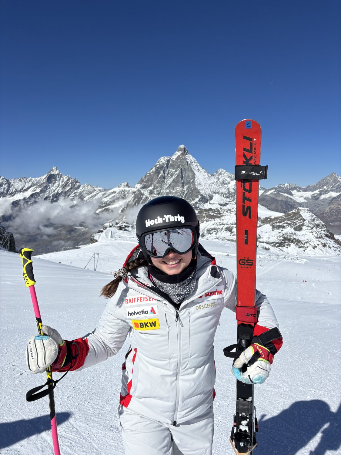 Allegra Frei, im weißen Anzug und mit Helm, steht lächelnd auf einem verschneiten Berghang, in der Hand Skistöcke und einen roten Ski. Im Hintergrund sind verschneite Gipfel und ein strahlend blauer Himmel zu sehen, hinter ihr das Matterhorn.