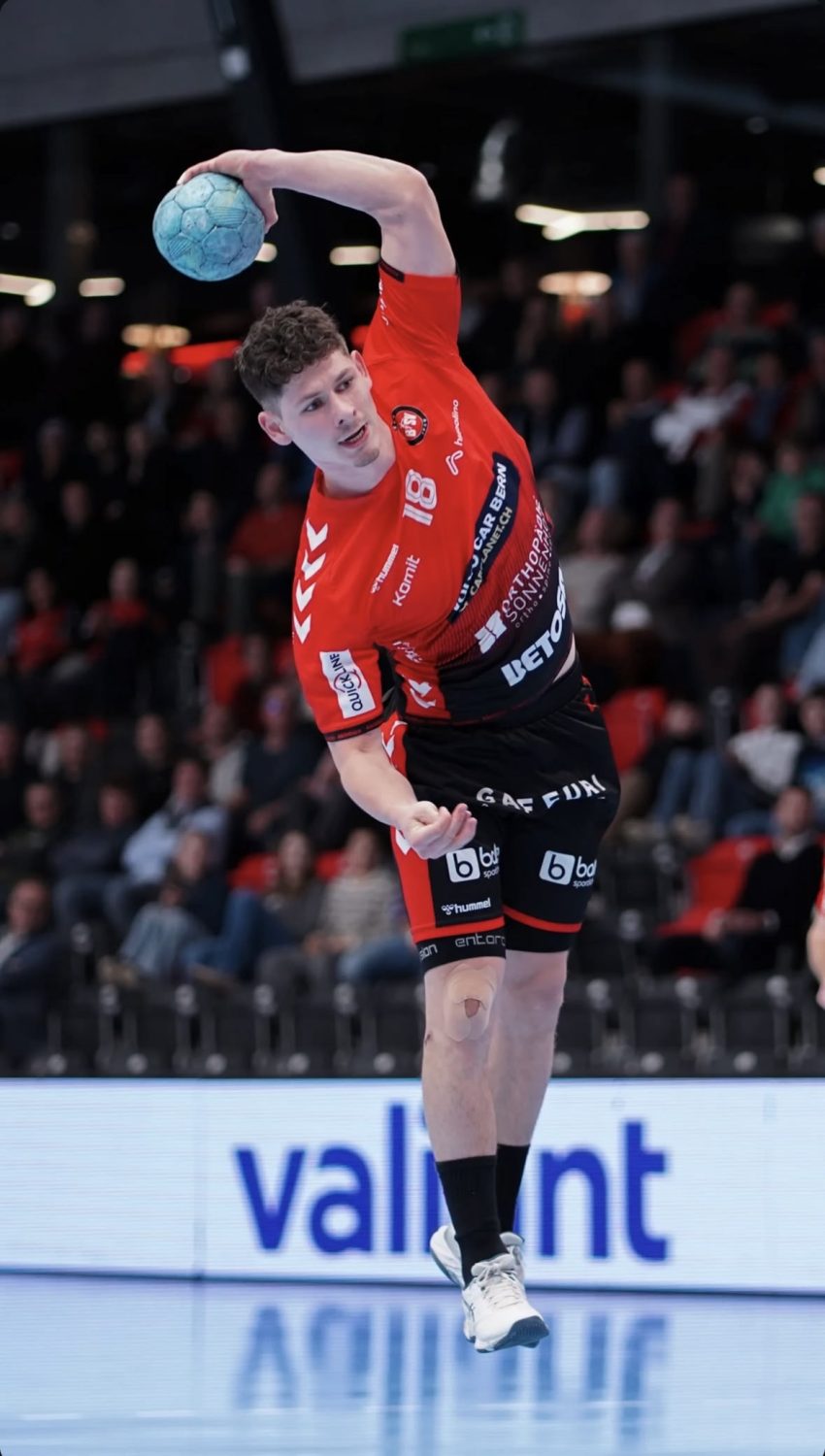 A male handball player in a red jersey is mid-air, about to throw a blue ball. He is focused, with spectators watching in the background. The scene takes place in an indoor sports arena.