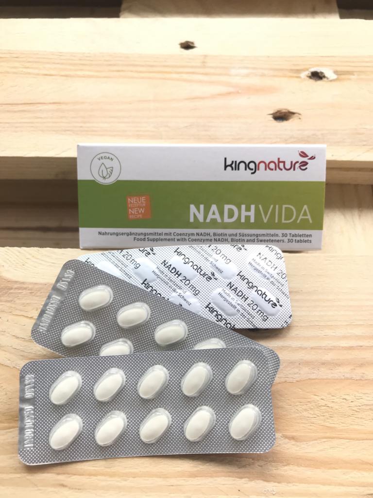 Hinter zwei Blisterpackungen mit weißen ovalen Tabletten auf hellem Holzgrund steht eine Schachtel mit der Aufschrift „kingnature NADH Vida“. Die Verpackung kennzeichnet das Produkt als vegan, neu und mit NADH und Q10.