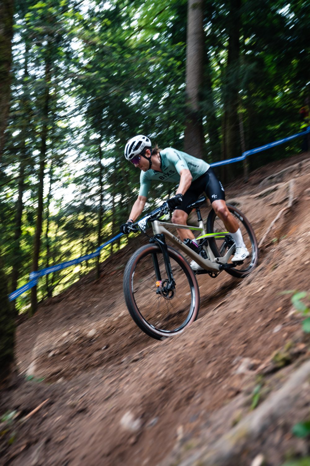 Ein Radfahrer, der einen Helm, eine Sonnenbrille und ein grünes Trikot trägt, fährt mit seinem Mountainbike einen steilen, unbefestigten Weg in einem Wald hinunter, der von Bäumen umgeben und durch ein blaues Streckenband gekennzeichnet ist.