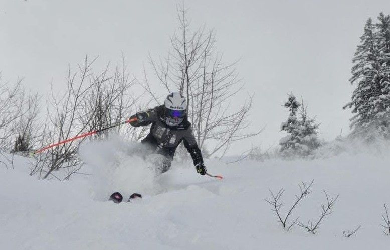 Allegra Frei, mit weissem Helm und Brille, carvt durch den frischen Pulverschnee an einem Hang, umgeben von blattlosen Sträuchern und schneebedeckten Bäumen unter einem wolkenverhangenen Himmel.