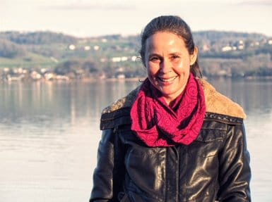 Eine Frau in einer schwarzen Lederjacke und einem roten Schal steht lächelnd neben einem ruhigen See, im Hintergrund sind grüne Hügel und Häuser zu sehen – ein Bild, das die friedliche Atmosphäre von Bettina Neuhaus‘ Praxis für TCM widerspiegelt.