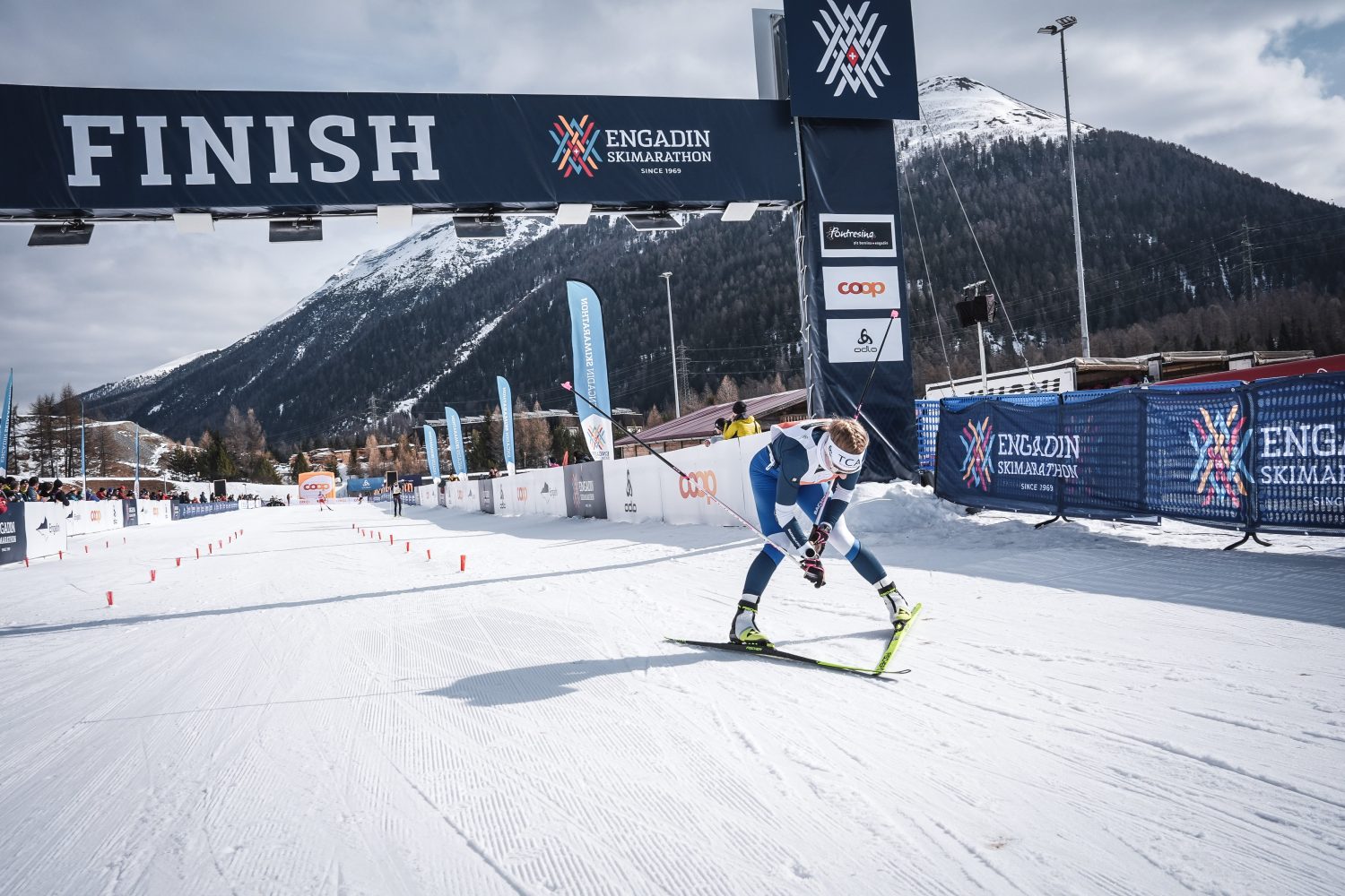 Lisa Naegeli überquert im Renndress die verschneite Ziellinie des Engadin Skimarathons. Im Hintergrund erheben sich die Berge, hinter den Absperrungen schauen Zuschauer zu.