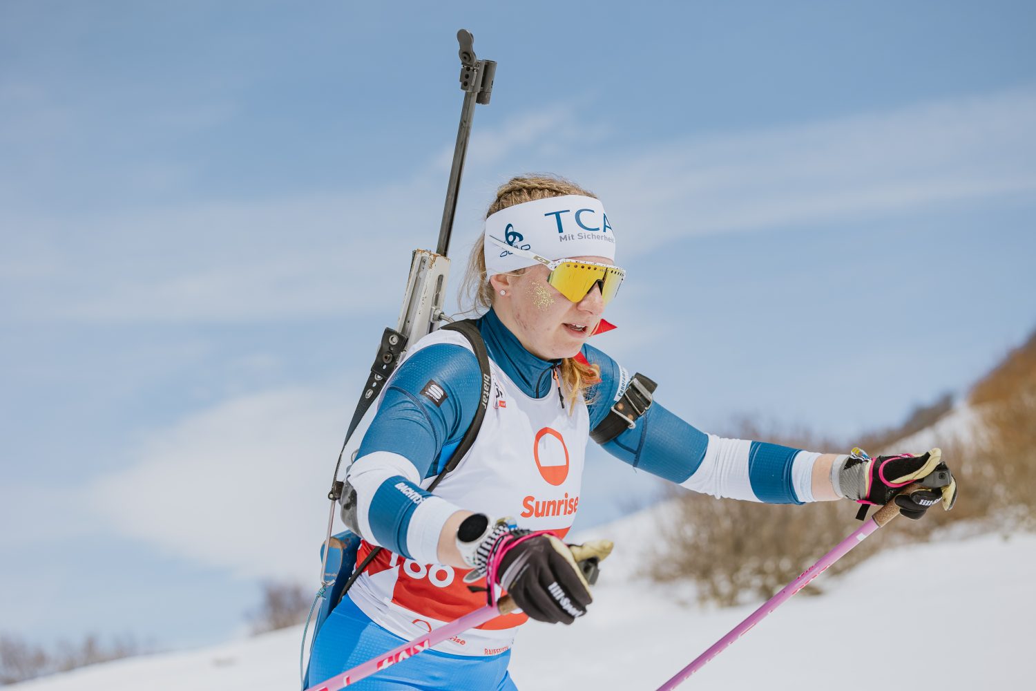 Lisa Naegeli, eine Biathletin in Wintersportkleidung, fährt mit einem Gewehr auf dem Rücken im Freien Ski. Sie trägt eine Sonnenbrille, ein weißes Stirnband und eine weiße Startnummer mit Sunrise und der Nummer 108. Im Hintergrund sind Schnee und Bäume zu sehen.