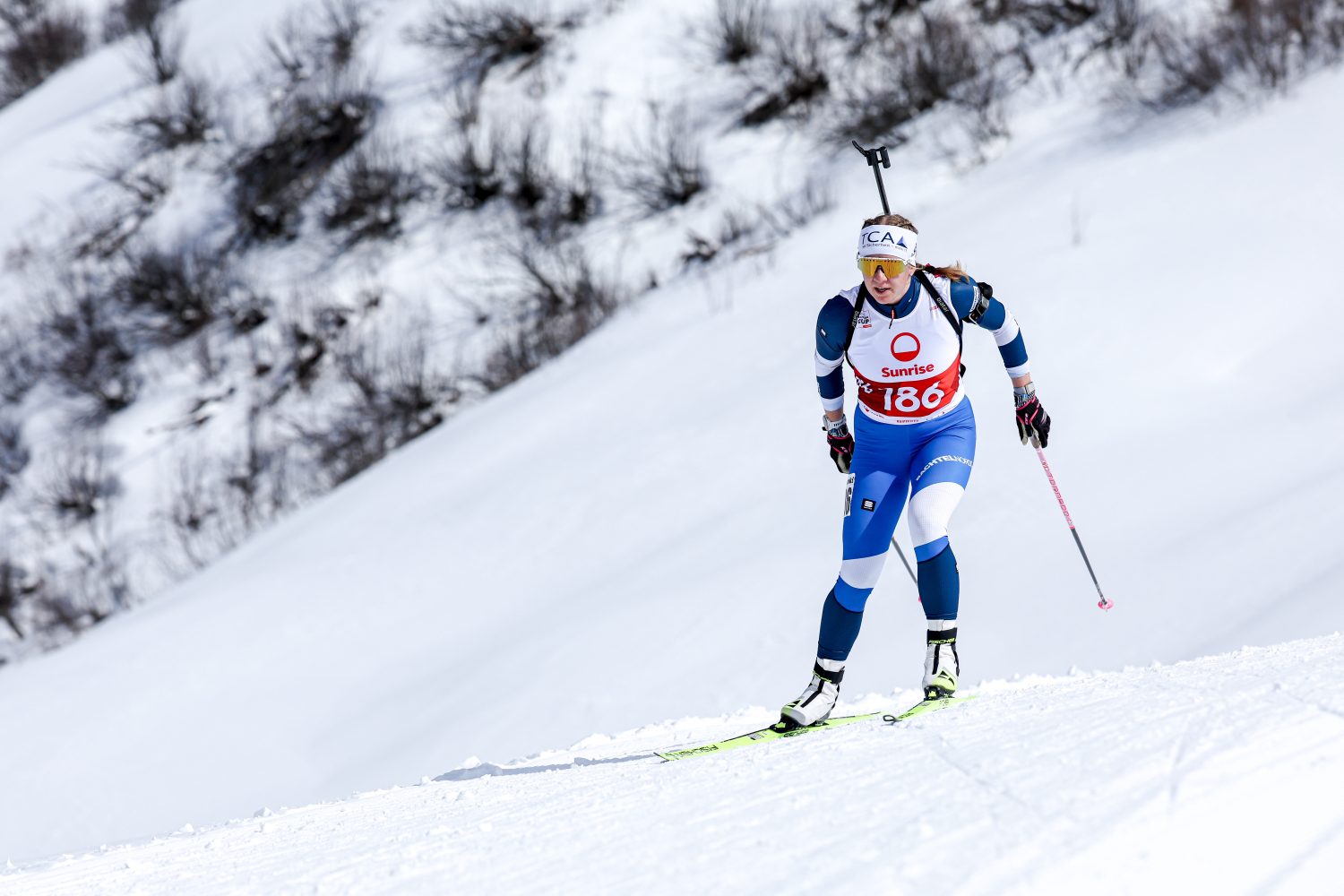 Die Biathletin Lisa Naegeli, die eine rote Startnummer 186 trägt, fährt auf einer verschneiten Piste mit einem Gewehr auf dem Rücken bergauf. Sie ist in einen blau-weißen Anzug gekleidet und benutzt Skistöcke, im Hintergrund sind schneebedeckte Sträucher zu sehen.
