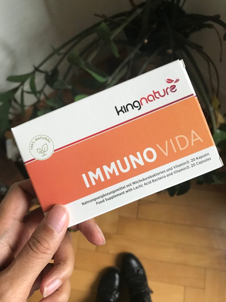 Eine Hand hält eine Packung kingnature IMMUNOVIDA Nahrungsergänzungsmittel mit Milchsäurebakterien und Vitamin D zur Stärkung des Immunsystems. Die orange-weiße Packung trägt ein grünes 100%-Natur-Etikett. Im Hintergrund sind Holzboden und Schuhe zu sehen.