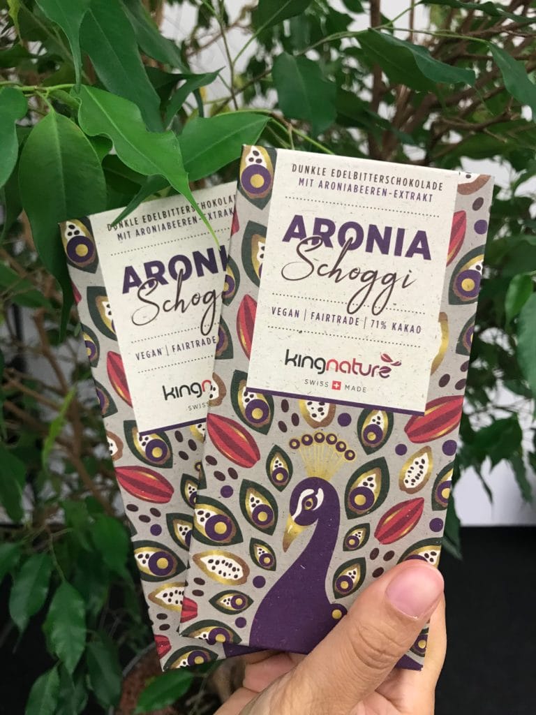 Eine Hand hält zwei Tafeln Kingnature Aronia Schokolade mit Kakaoanteil 71 % und buntem Pfauendesign auf der Verpackung, vor grünen Blattpflanzen – perfekt für gesunden Schoggi-Genuss.