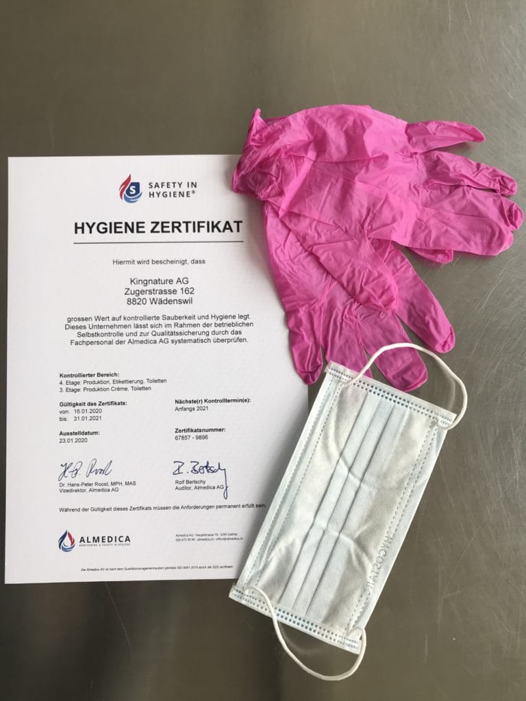 Auf einem Tisch liegt ein Hygienezertifikat in deutscher Sprache aus, daneben ein Paar rosafarbene Einweghandschuhe, eine blaue OP-Maske und Sicherheitsausrüstung.