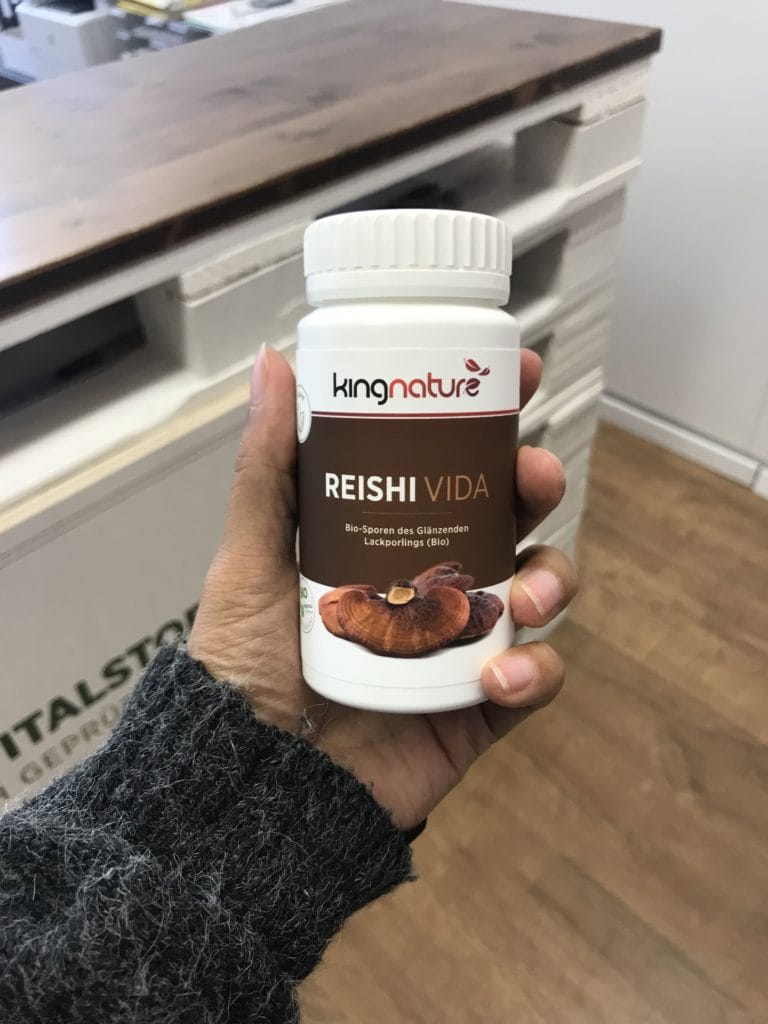 Eine Hand hält eine weiße Flasche Reishi Vida, ein neues Produkt von kingnature, mit Reishi-Pilzen auf dem Etikett. Im Hintergrund sind eine Holztheke und ein Teil eines weißen Schranks zu sehen.
