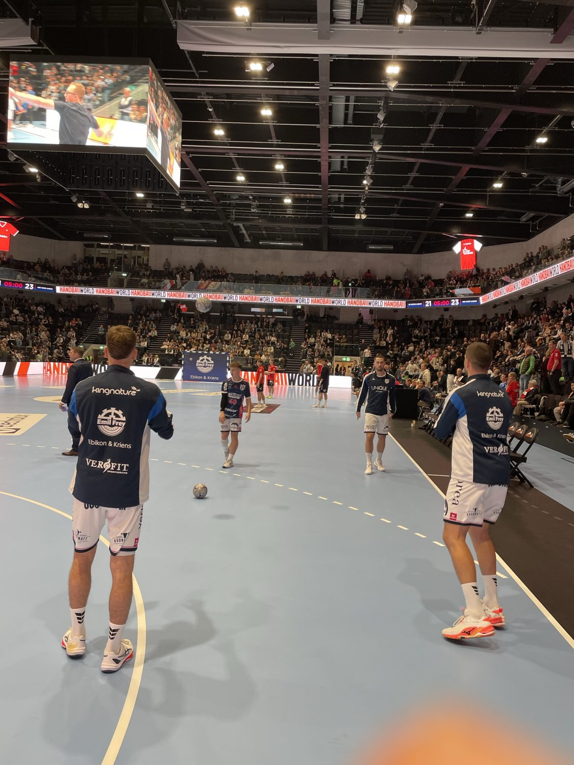 Vier Handballer des HC Kriens-Luzern in blau-weißen Trikots wärmen sich auf einem Hallenplatz auf, während die Zuschauer auf der Tribüne zusehen. Helle Oberlichter erhellen die Halle in der Schweiz, in der ein Spiel beginnen soll.