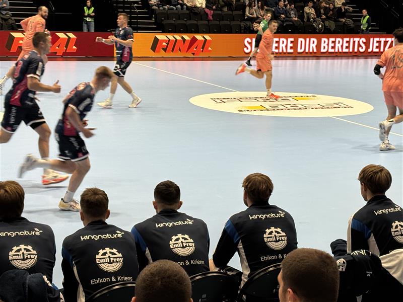 Spieler in blauen und rosafarbenen Trikots des HC Kriens-Luzern sitzen auf der Bank und beobachten ihre Mannschaftskameraden auf dem Spielfeld während eines Handballspiels. Im Vordergrund spielen die Gegner in Orange, im Hintergrund sind FINAL und eine deutsche Fahne zu sehen.