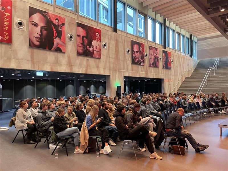 Eine große Gruppe von Menschen sitzt auf Stühlen in einem modernen Innenraum und blickt nach vorne. Über ihnen hängen Porträts mit Sportmotiven - einige davon zum Thema Handball und HC Kriens-Luzern - und rechts ist eine breite Treppe zu sehen, die die lebendige Sportkultur der Schweiz widerspiegelt.