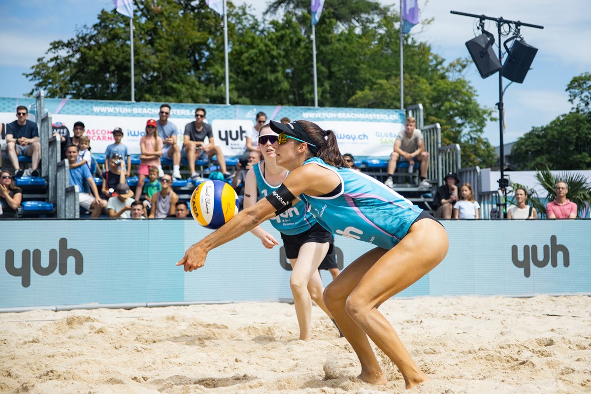 Janick Schaltegger und ein Teamkollege treten in Sportuniformen und Sonnenbrillen auf einem Sandplatz im Beachvolleyball an, während Fans von der Tribüne aus zuschauen und im Hintergrund Bäume und Banner zu sehen sind.