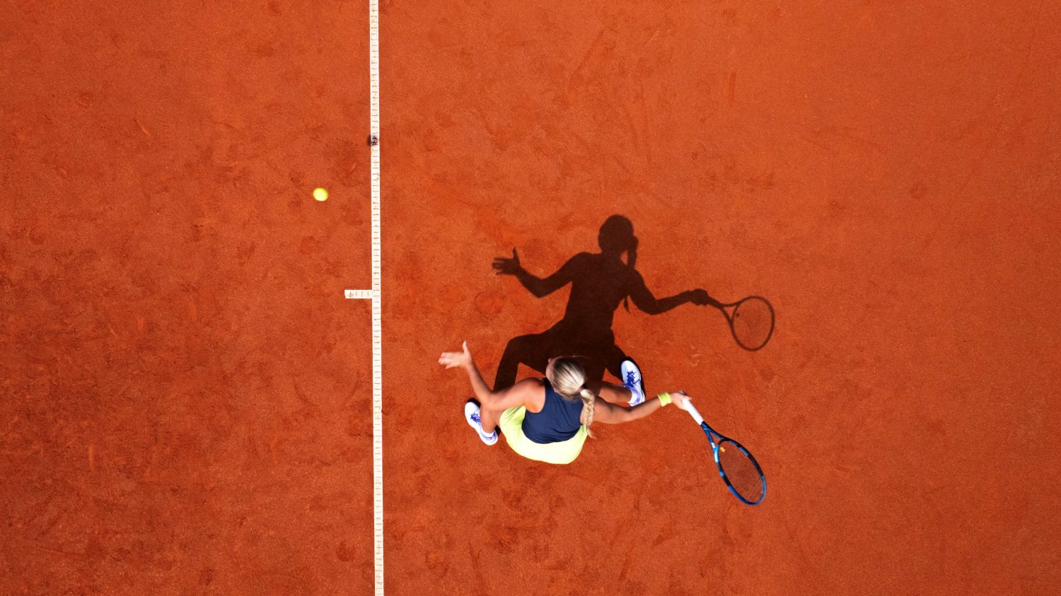 Luftaufnahme eines Tennisspielers auf einem Sandplatz, der sich darauf vorbereitet, einen Ball mit dem Schläger nahe der Grundlinie zu schlagen. Der Schatten des Spielers ist auf der Platzoberfläche sichtbar.