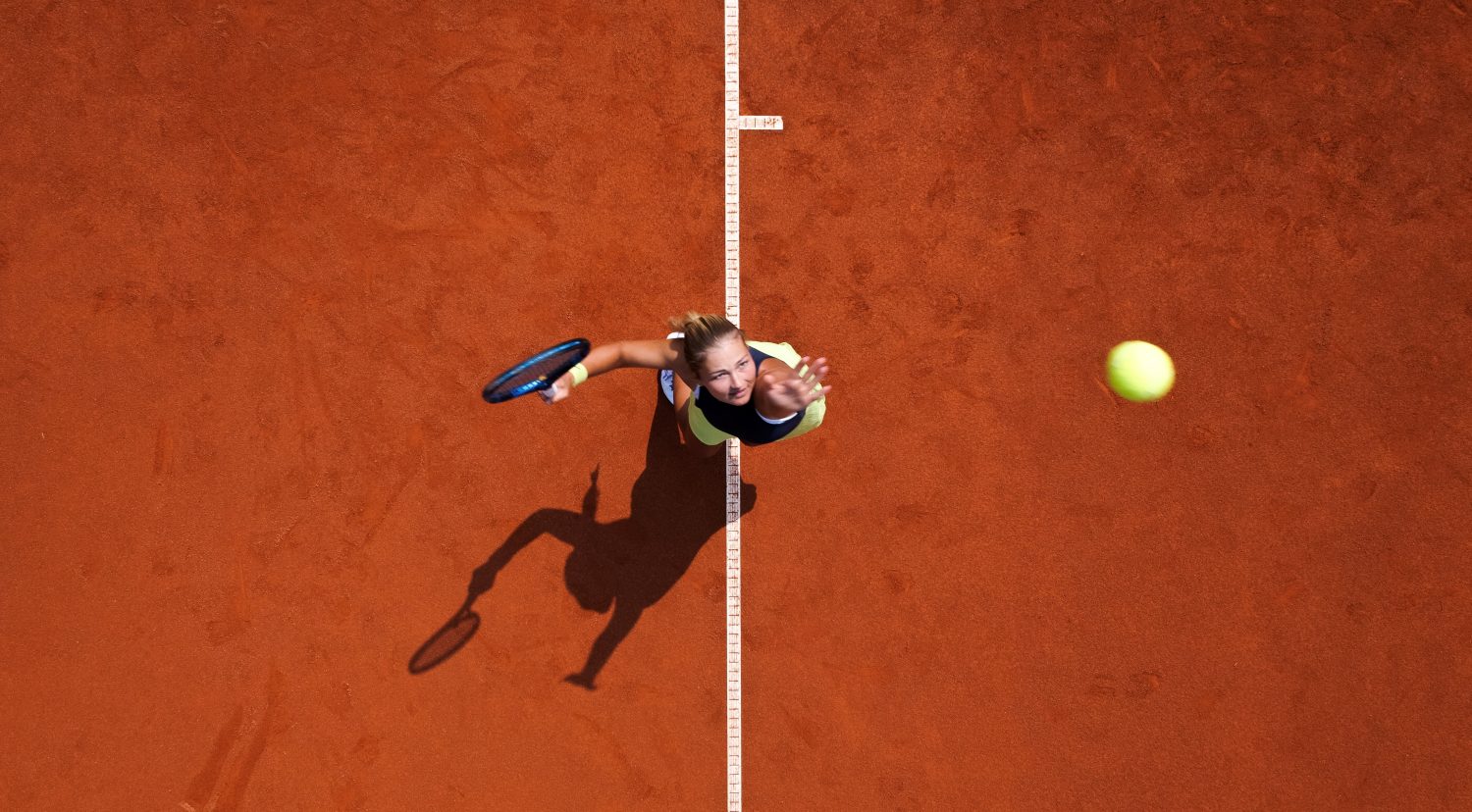 Luftaufnahme eines Tennisspielers beim Aufschlag auf einem Sandplatz. Der Ball befindet sich in der Luft und der Schatten des Spielers ist auf dem Boden sichtbar.