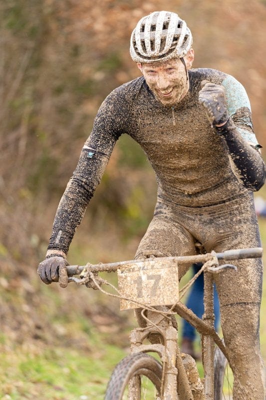 Johannes Közle, mit Schlamm bedeckt, lächelt und reckt jubelnd die Faust, während er während des Rennens mit einem Mountainbike auf einem Feldweg fährt und dabei einen Helm und Sportkleidung von Kingnature trägt.