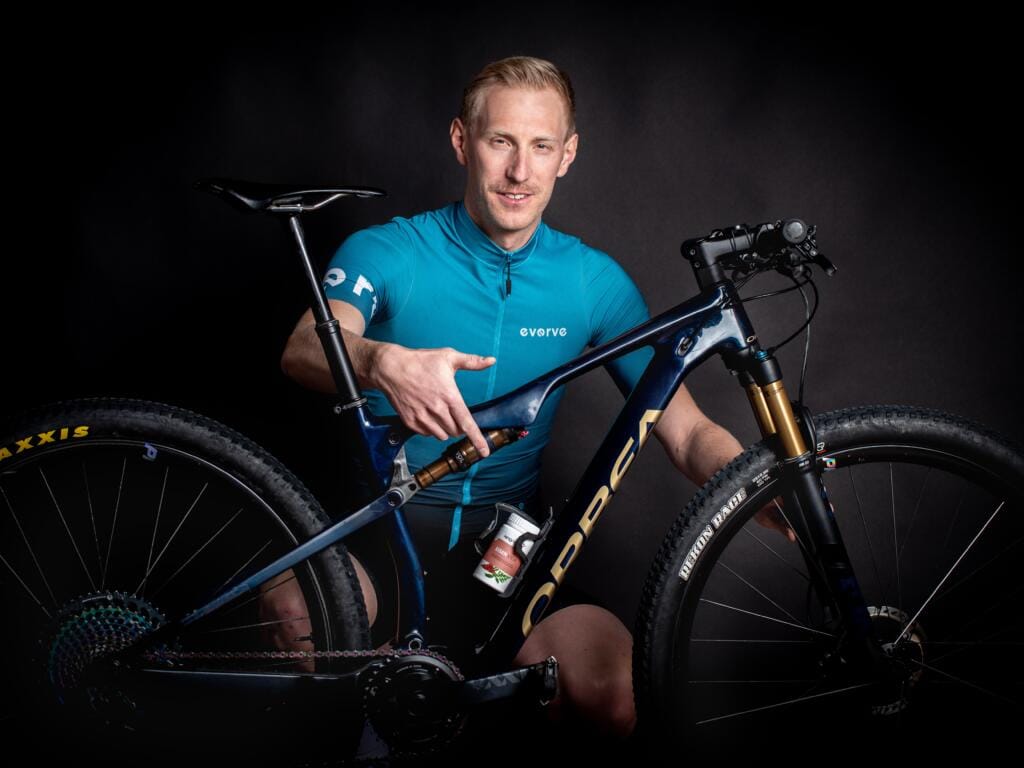 Ein Mann in einem blauen Radtrikot sitzt da, hält ein dunkelblaues Mountainbike in der Hand und zeigt auf den Rahmen. Der schwarze Hintergrund hebt sowohl den Radfahrer als auch sein Mountainbike hervor.