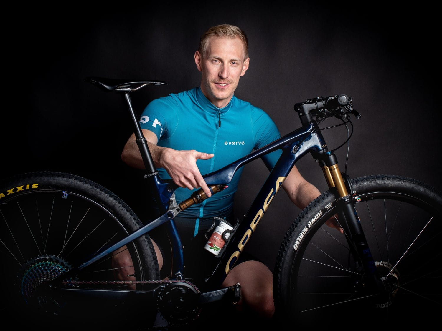 Ein Mann in einem blauen Radtrikot sitzt da, hält ein dunkelblaues Mountainbike in der Hand und zeigt auf den Rahmen. Der schwarze Hintergrund hebt sowohl den Radfahrer als auch sein Mountainbike hervor.