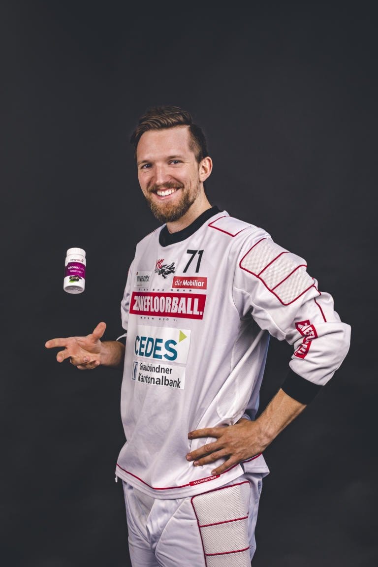 Ein lächelnder Mann in weißer Sportuniform steht vor einem schwarzen Hintergrund und wirft mit einer Hand eine kleine weiße Nahrungsergänzungsflasche in die Luft, während die andere auf seiner Hüfte ruht.