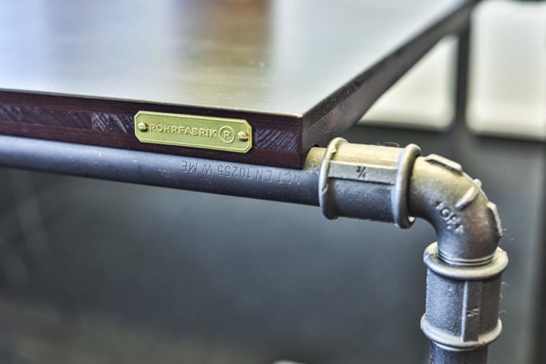 Nahaufnahme einer Tischecke mit dunkler Holzplatte, Beinen und Beschlägen aus Metallrohren im Industriestil und einem Metalletikett mit der Aufschrift „ROHRFABRIK“ – eine ideale Ergänzung für jede Büromöbel-Kollektion.