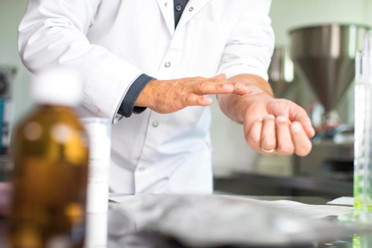 Eine Person in einem weißen Laborkittel reibt ihr linkes Handgelenk mit der rechten Hand in einer Laborumgebung, in der verschwommene Flaschen und Geräte sichtbar sind. Die Szene vermittelt den Eindruck eines dermatologisch getesteten Produkts.