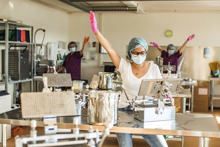 Eine Frau mit Maske, Haarnetz und rosa Handschuhen hebt ihren Arm, während sie in einer laborähnlichen Umgebung arbeitet. Zwei weitere maskierte Personen gesellen sich zu ihr. Sie wirken alle fröhlich und verspielt inmitten von Edelstahlgeräten – und bringen gute Nachrichten vom kingnature-Team.