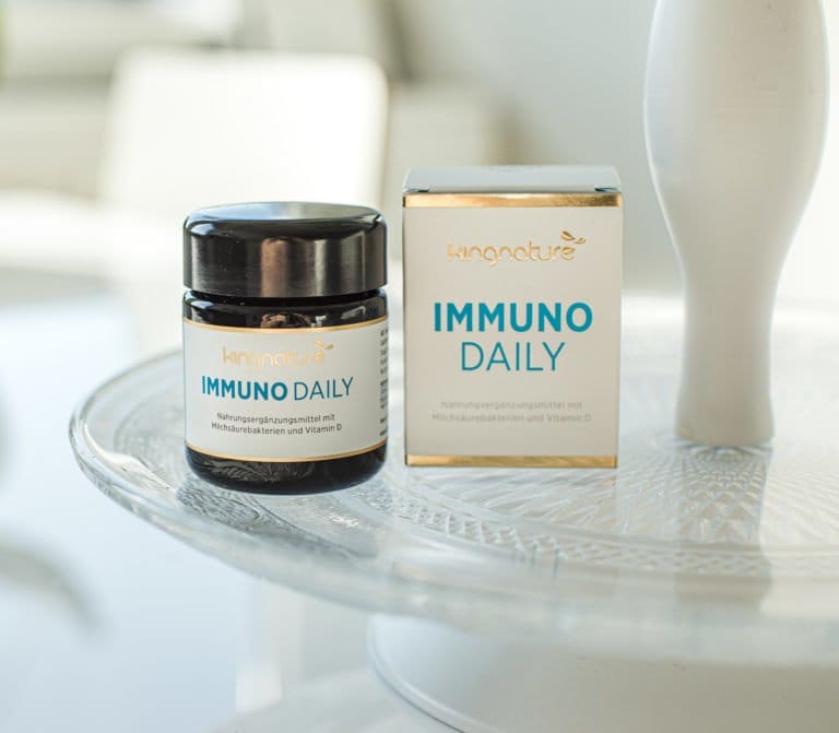Ein Glas und eine Schachtel Immuno Daily, ein neues Produkt, stehen auf einem weißen Glastisch. Beide Verpackungen sind mit blauem und goldenem Text und Markenzeichen versehen, der Hintergrund ist von sanftem Tageslicht erhellt.