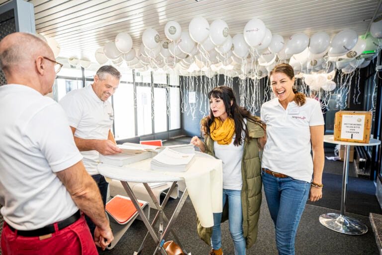 Vier Personen stehen lächelnd und unterhalten sich mit Papieren um einen Tisch. Sie befinden sich in einem Raum, dessen Decke mit vielen weißen Luftballons geschmückt ist, und feiern vermutlich ihren Erfolg am Tag der offenen Tür. Drei tragen weiße Hemden mit Namensschildern; einer trägt einen gelben Schal und einen grünen Mantel.