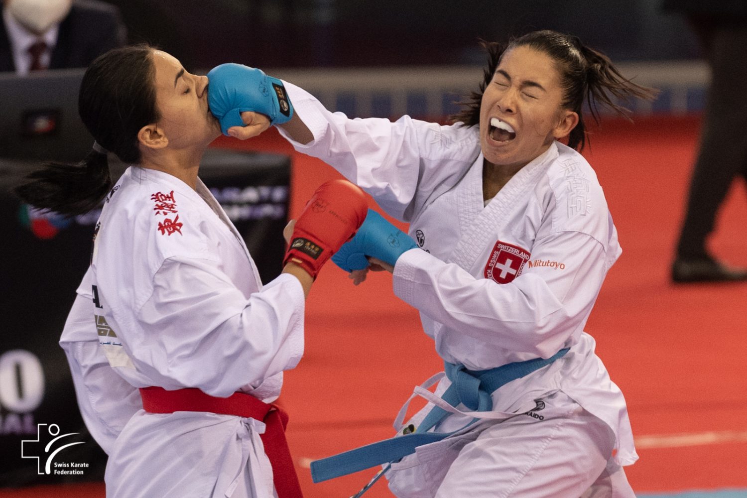 Two female karate athletes in white uniforms compete; one wearing red gloves is struck in the face by the other, who wears blue gloves. Both show intense expressions during the match on a red mat.
