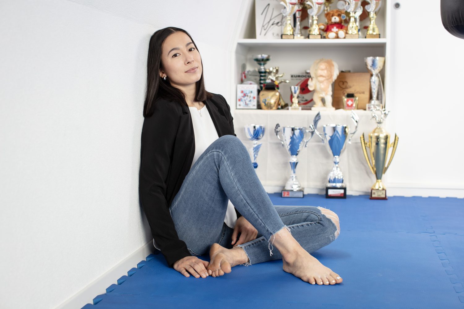 A young woman sits barefoot on a blue mat, leaning against a white wall. Behind her is a shelf displaying numerous trophies, medals, and awards. She wears jeans, a white top, and a black jacket, looking calmly at the camera.
