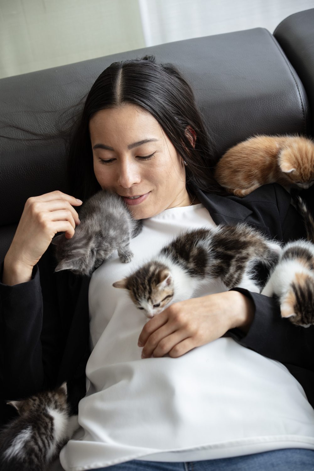 A woman lying on a couch smiles gently while several small kittens climb on her chest and arm. The scene appears cozy and affectionate.