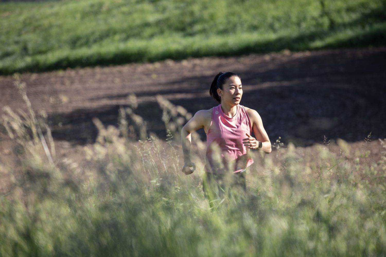A woman wearing a pink sleeveless top jogs outdoors through tall grass, with a field and greenery in the background on a sunny day.