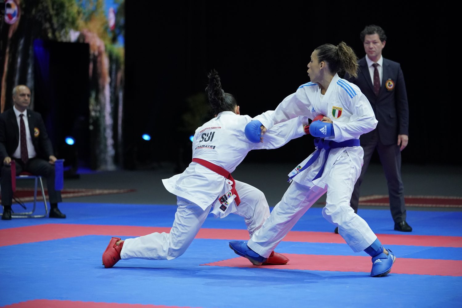 Two female karate athletes compete in a match on a blue and red mat. One is punching while the other leans forward to counter. Both wear white uniforms with colored belts and gloves. Judges sit and stand in the background.