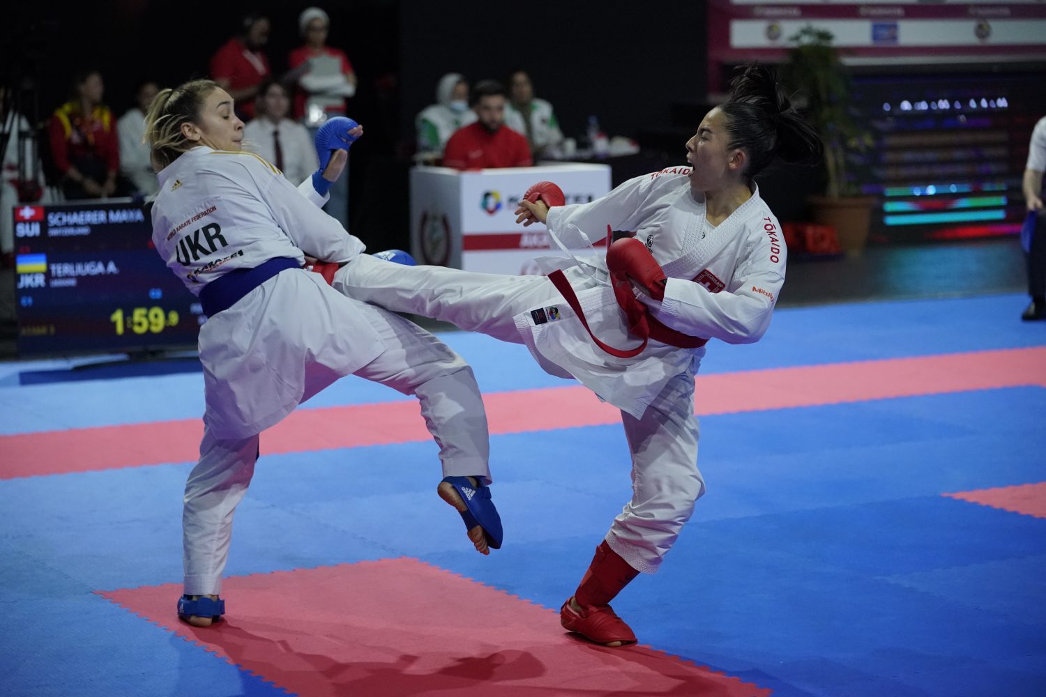 Two female karate athletes spar in a competition. The athlete on the right, wearing red gloves and gear, delivers a high kick to her opponent, who wears blue gloves and gear. Both are dressed in white karate uniforms on a blue and red mat.