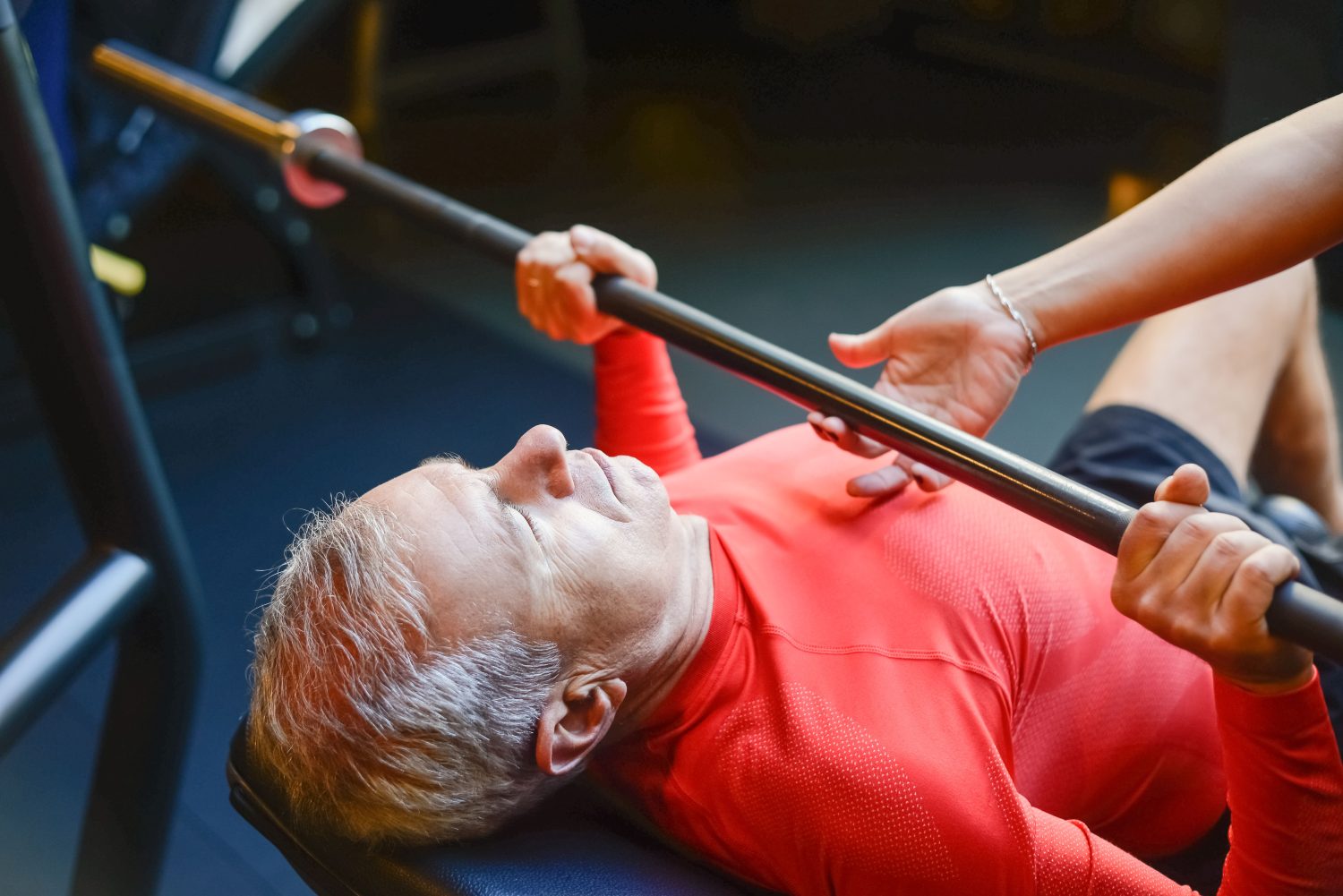 Ein älterer Mann in einem roten Hemd liegt auf einer Bank und hält eine Hantelstange fest, während ihm eine andere Person beim Bankdrücken im Fitnessstudio mit den Händen hilft – ein Beispiel dafür, wie gezieltes Training dazu beitragen kann, den Muskelabbau im Alter zu stoppen und der Sarkopenie entgegenzuwirken.