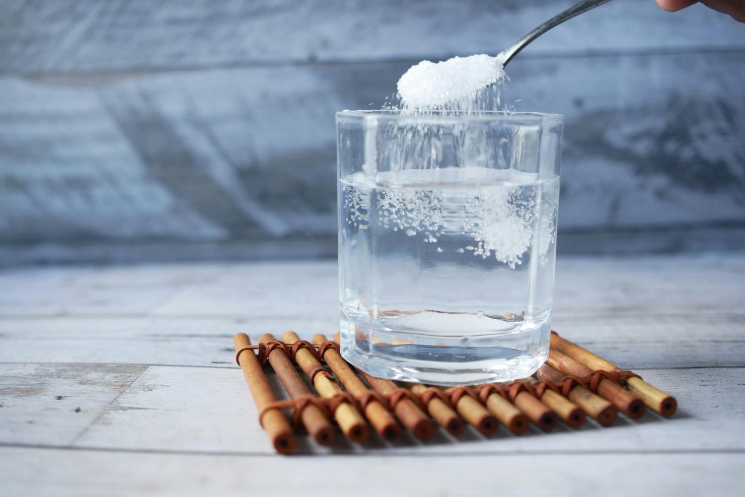 Ein Löffel weißes Pulver, möglicherweise Kreatin, das in ein Glas mit klarem Wasser gegeben wird, das auf einer Bambusmatte vor einem hellen Holzhintergrund ruht - der Moment, bevor seine Wirkung und Dosierung einsetzt.